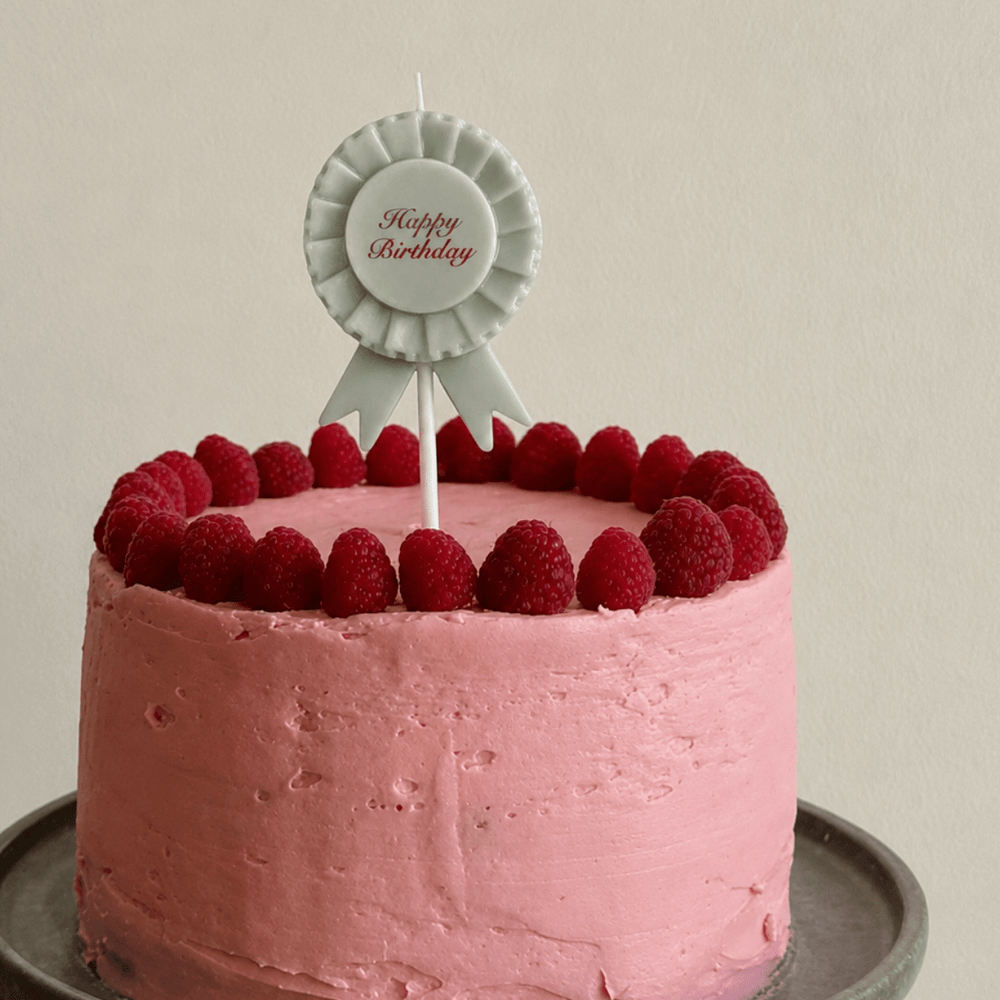 A round pink frosted cake topped with fresh raspberries and a white rosette-style 'Happy Birthday' candle, held on a metal cake stand against a plain light background.