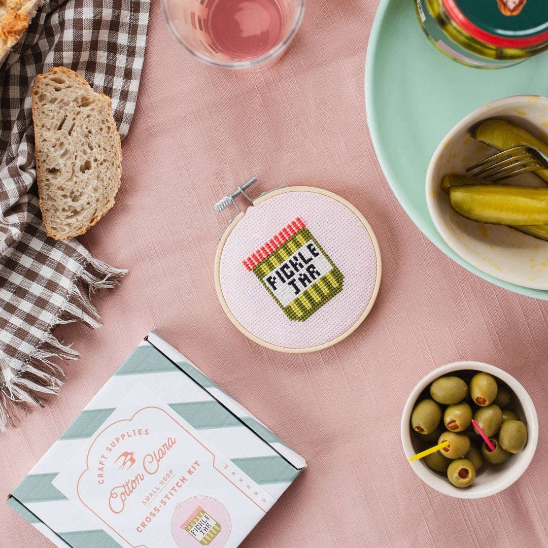 Pickle jar cross stitch hoop on a pink cloth with bread, pickles, and olives