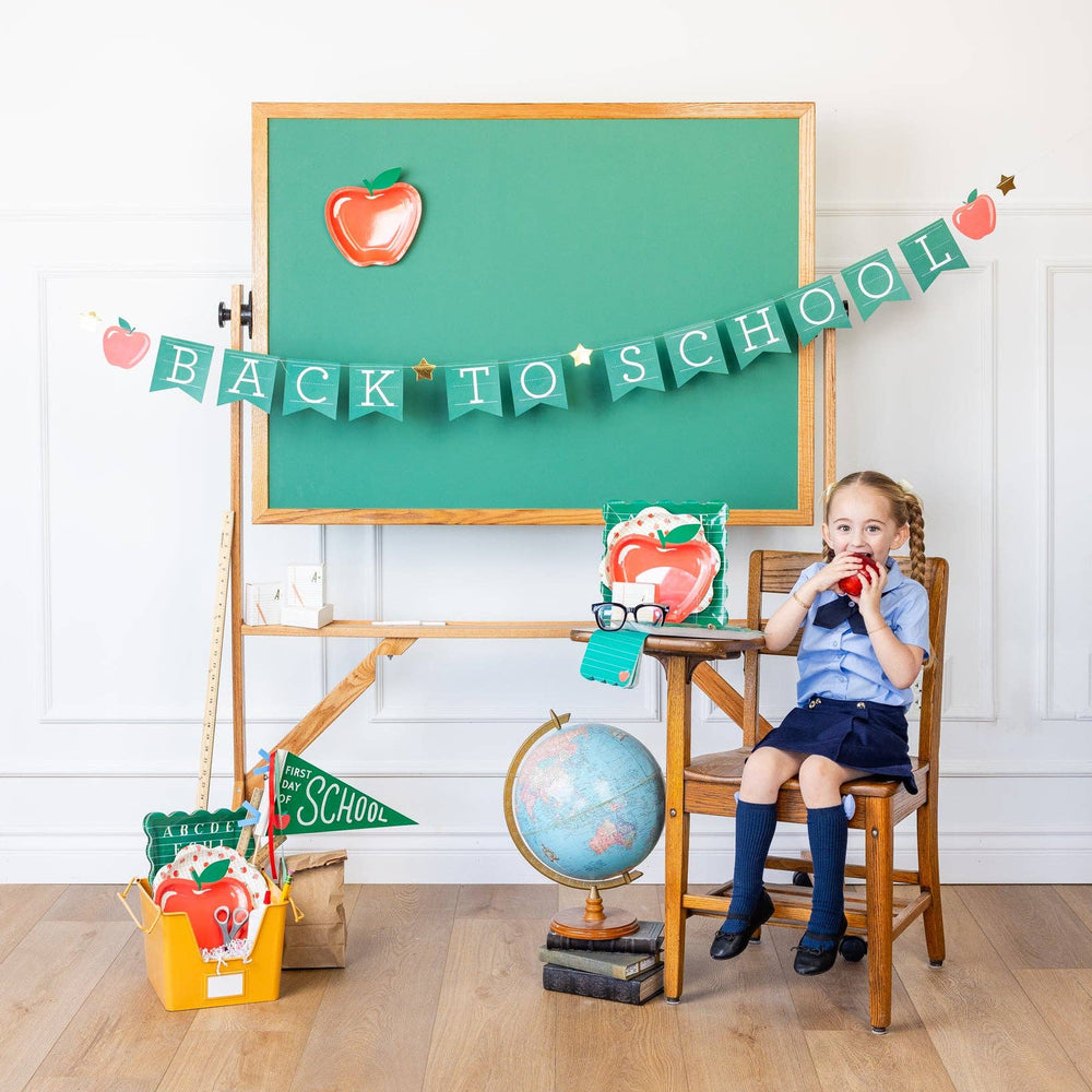 Girl in uniform by a chalkboard with the green Back to School banner hung above at a first day of school party