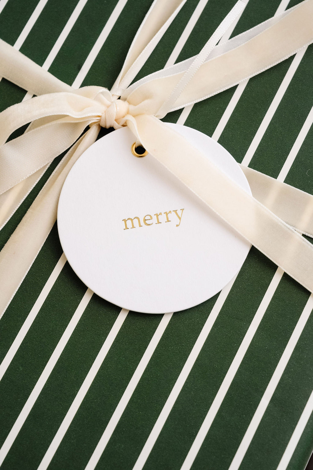 Round gold foil merry tag tied with cream velvet ribbon on green and white striped gift wrap