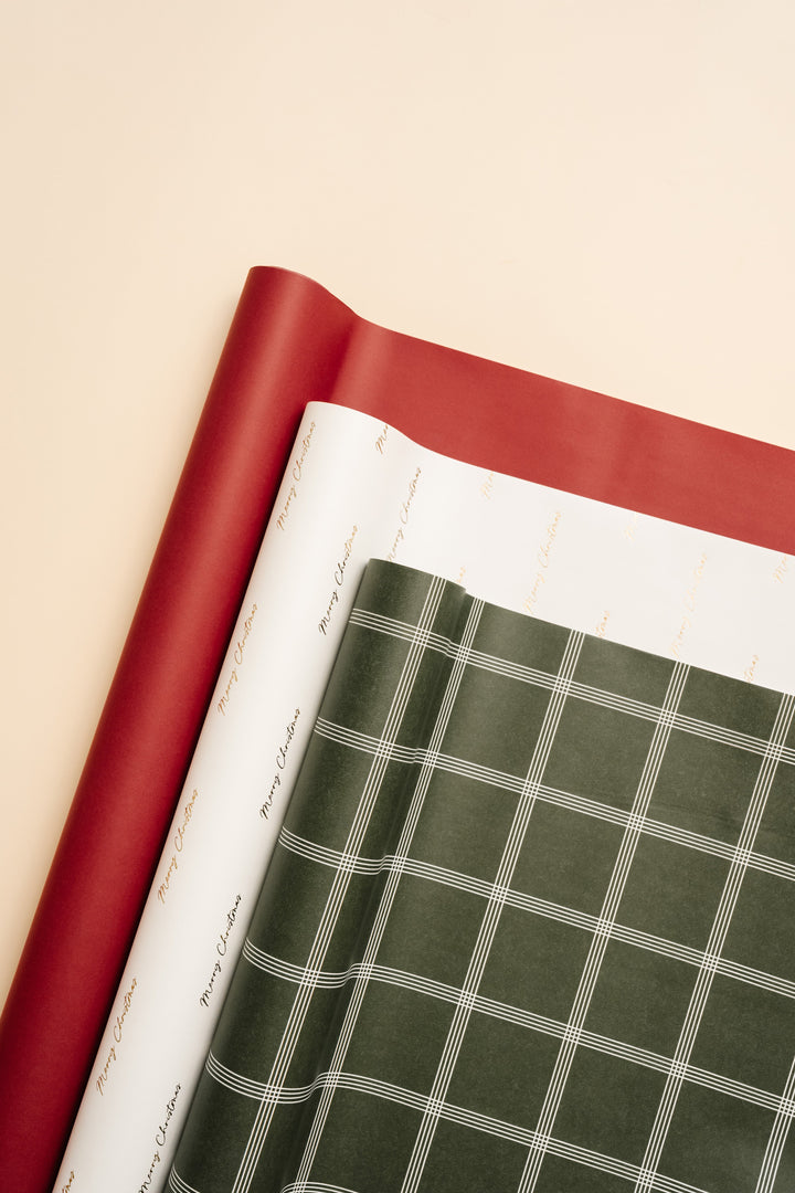 Layered sheets of solid red, gold script, and green plaid Christmas wrapping paper on cream