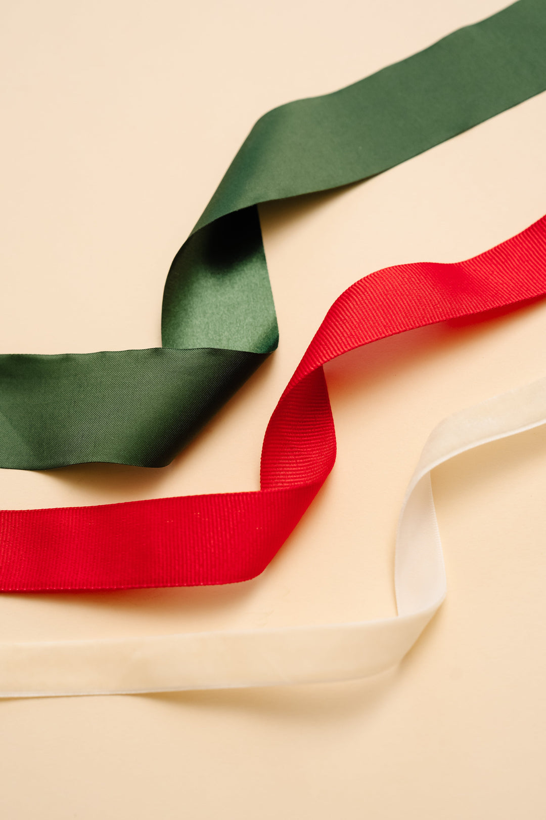 Green satin, red grosgrain, and cream velvet ribbon lengths curling across a cream backdrop