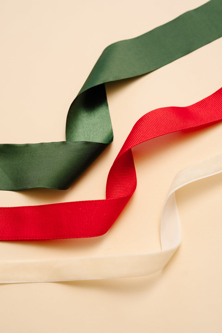 Green satin, red grosgrain, and cream velvet ribbon lengths curling across a cream backdrop