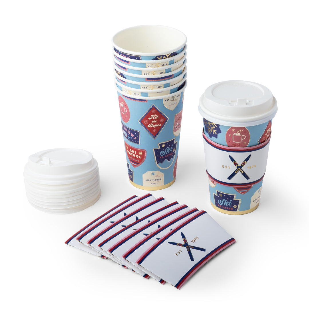 A set of custom paper coffee cup accessories on a white background, including a stack of paper cups with a light blue and red patch print, a stack of white paper lids, a stack of white sleeve wraps with a navy and red cross design, and a fan of small white napkins with red and navy stripes.