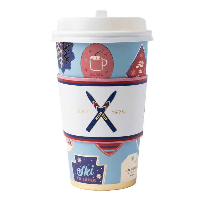 A reusable paper coffee cup with a white lid and a decorative wraparound sleeve. The cup features a light blue background with winter-themed graphics, including snowflakes, a mug with steam, a red triangle with white text, and a navy-and-red crossed-knife logo with the words 'EST.