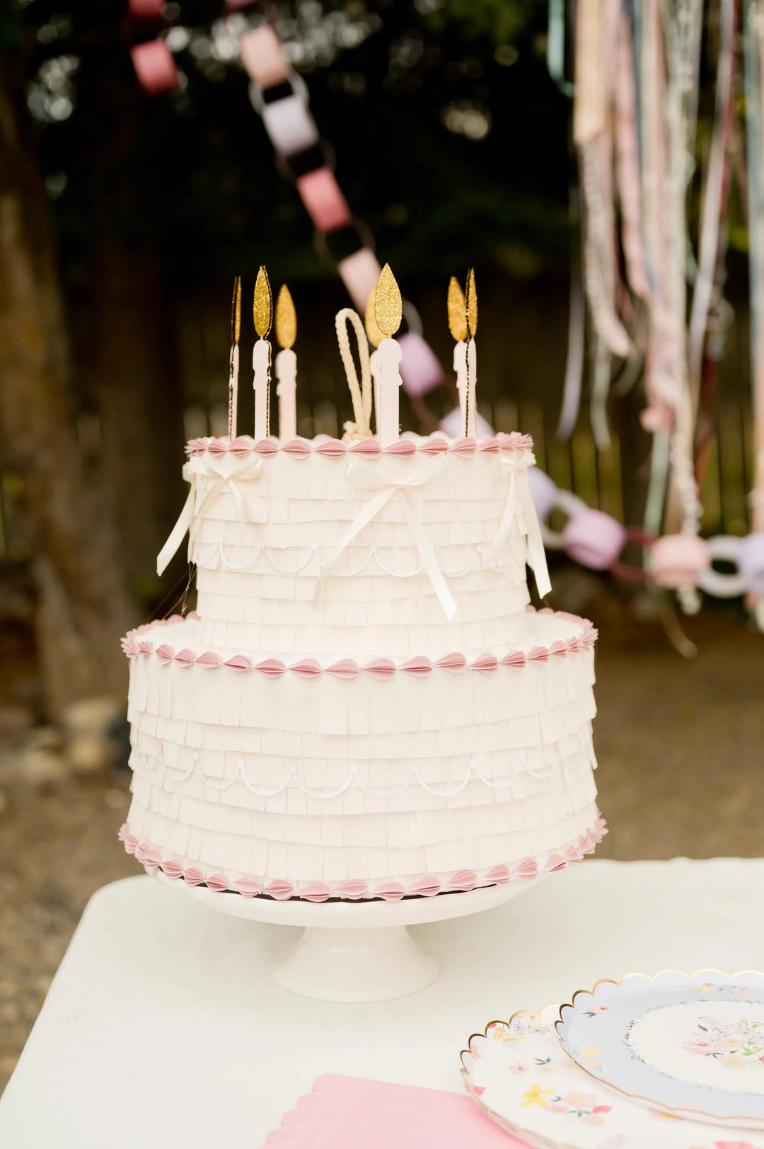 Two-tiered pinata cake with pink frosting and candles on a white stand, with decorative ribbons in the background.