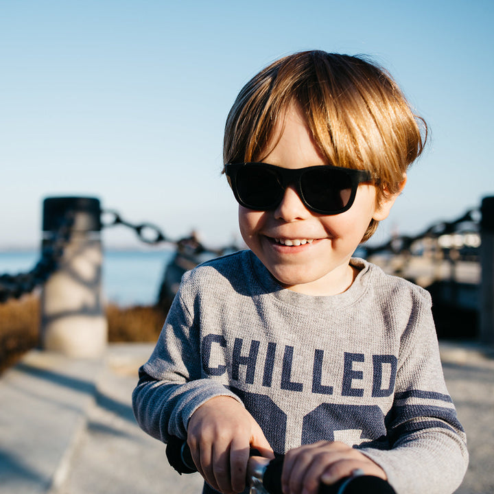 A smiling boy on a scooter wearing black aviator sunglasses outdoors by the water.