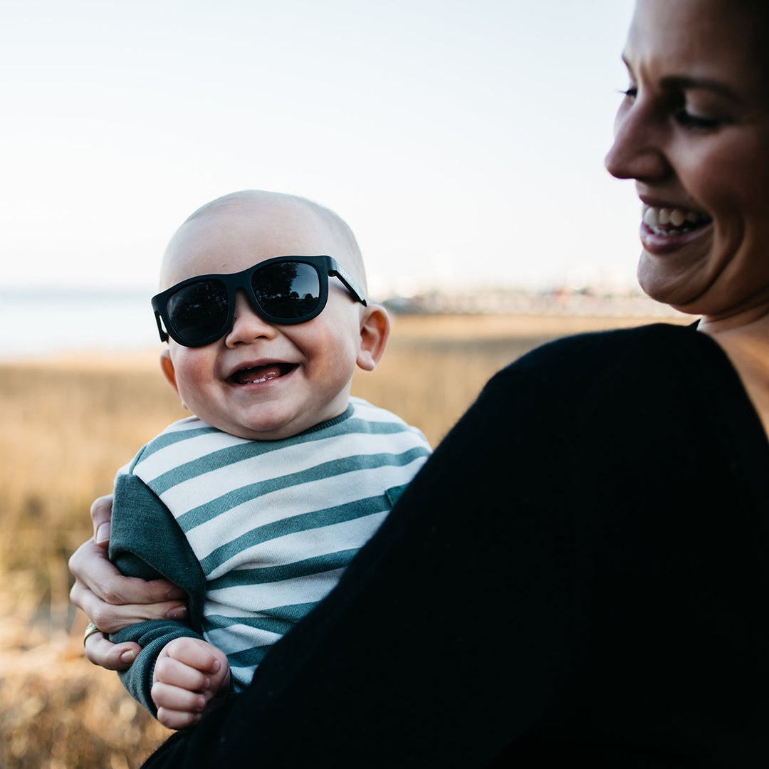 A laughing baby in a striped shirt wearing black aviator sunglasses while held by a smiling woman outdoors.