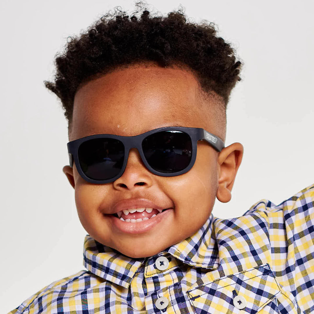 A smiling young boy in a yellow checkered shirt wearing black aviator sunglasses against a white backdrop.