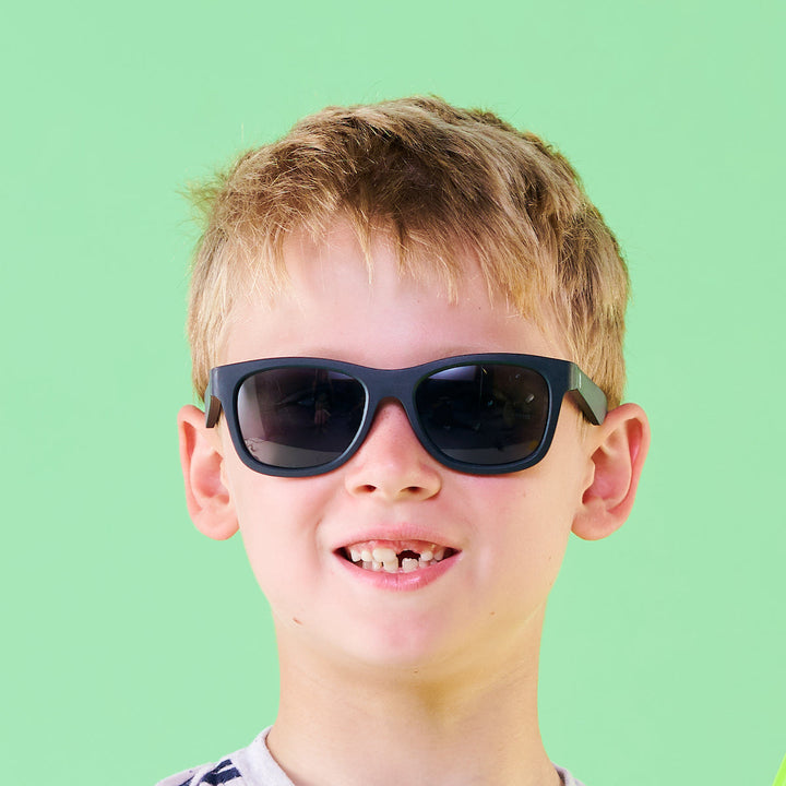 A smiling boy wearing black aviator sunglasses against a green backdrop.