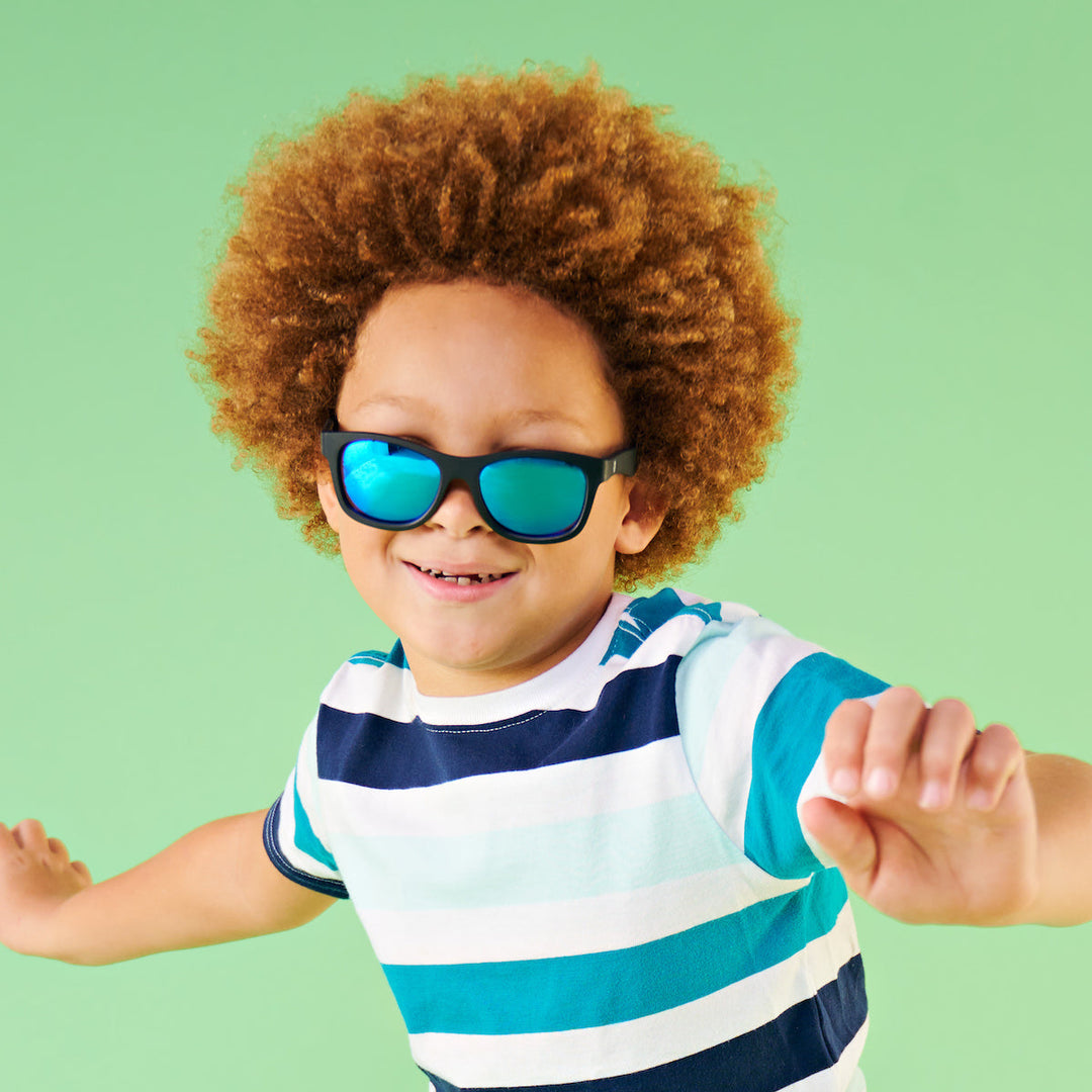 A boy in a striped shirt wearing black aviator sunglasses with blue mirrored lenses against a green backdrop.