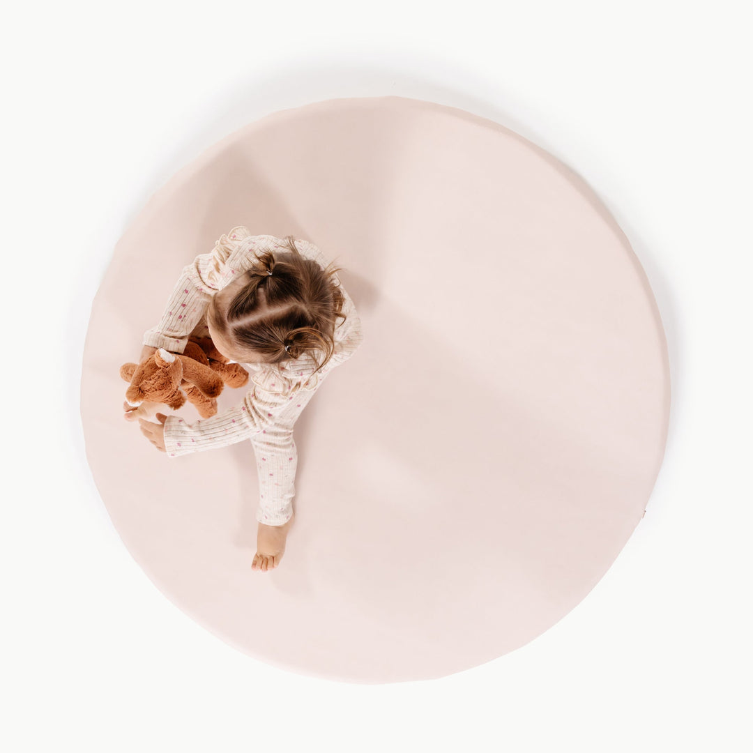 Cream blush round padded baby play mat, overhead view with a toddler and plush toy.