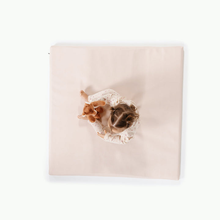Cream blush square padded baby play mat, overhead view with a toddler and plush toy.