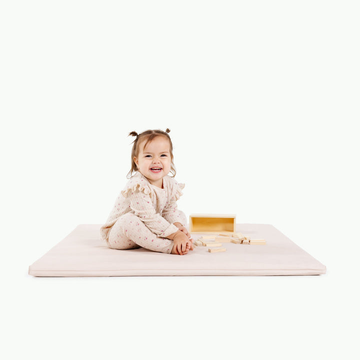 Cream blush padded baby play mat with a smiling toddler stacking wooden blocks.