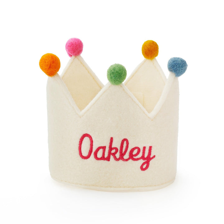Ivory felt crown with Oakley embroidered in red script and multicolor pom-poms, front view on white