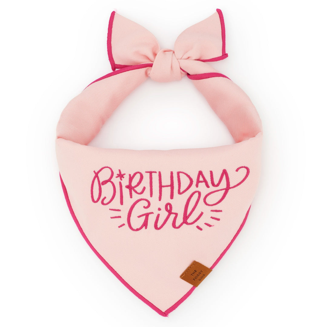 Light pink dog bandana embroidered Birthday Girl in hot pink script, tied flat on white