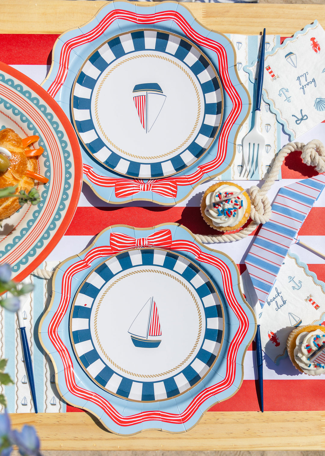 Overhead nautical place setting with striped sailboat plates, beach club napkins, and a cupcake on a red striped runner