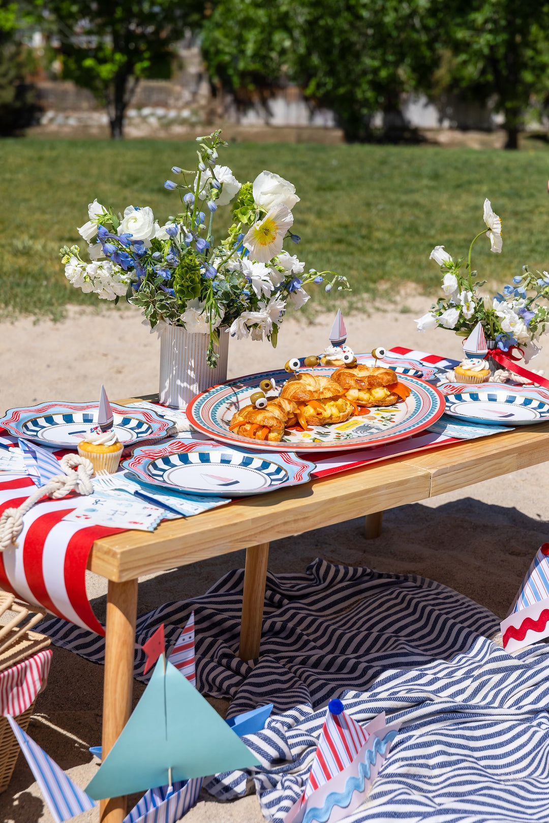 Nautical Fourth of July picnic table outdoors with striped sailboat plates, a striped runner, and croissant sailboat snacks