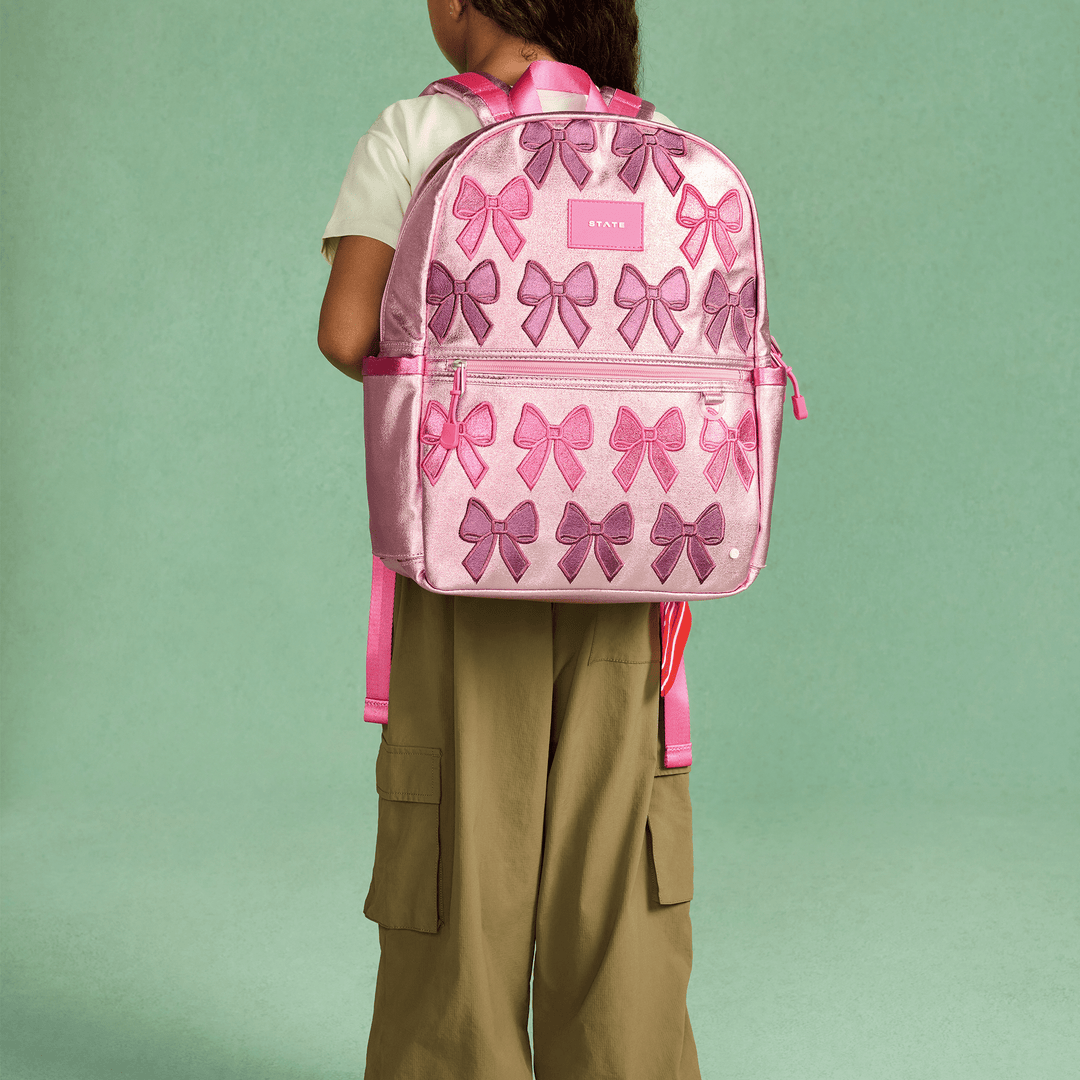 Child wearing the pink Patchwork Bows STATE Kane backpack against a green wall