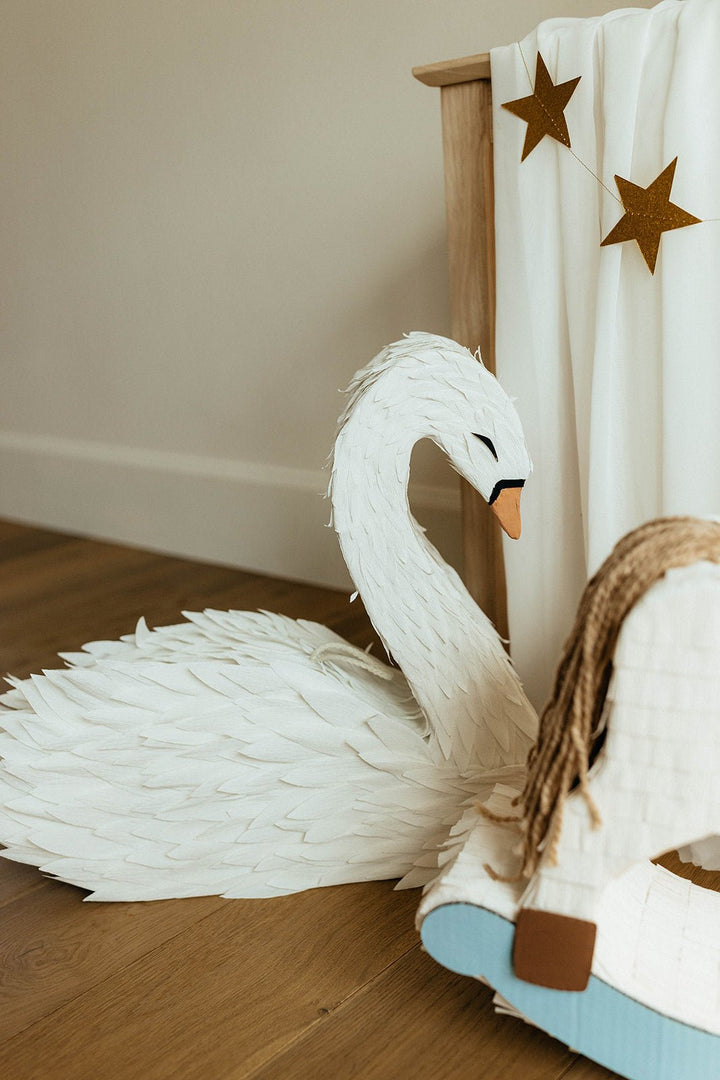 White swan pinata decoration on a wooden floor next to a bed with star-shaped decorations.