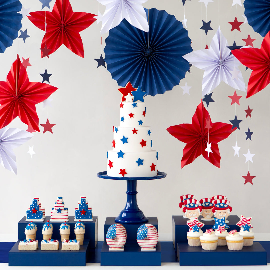 Decorative setup with a cake, cupcakes, and paper flowers in red, white, and blue on a patterned background.