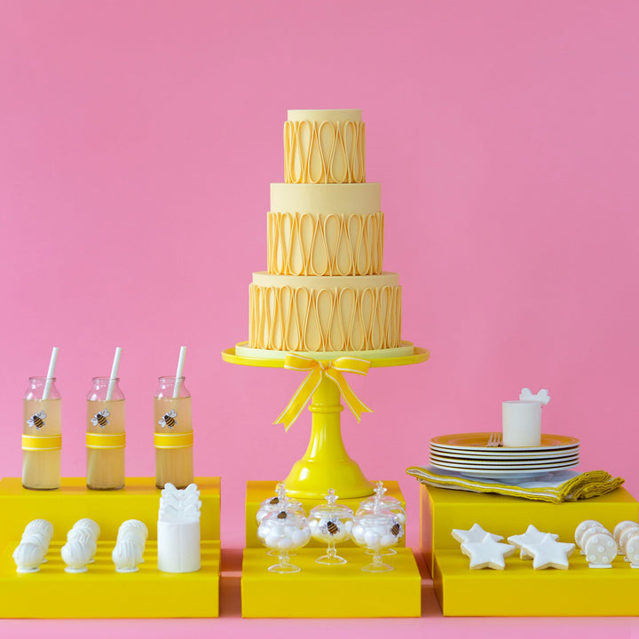 Three-tiered cake on a yellow stand with drinks and snacks on a pink background