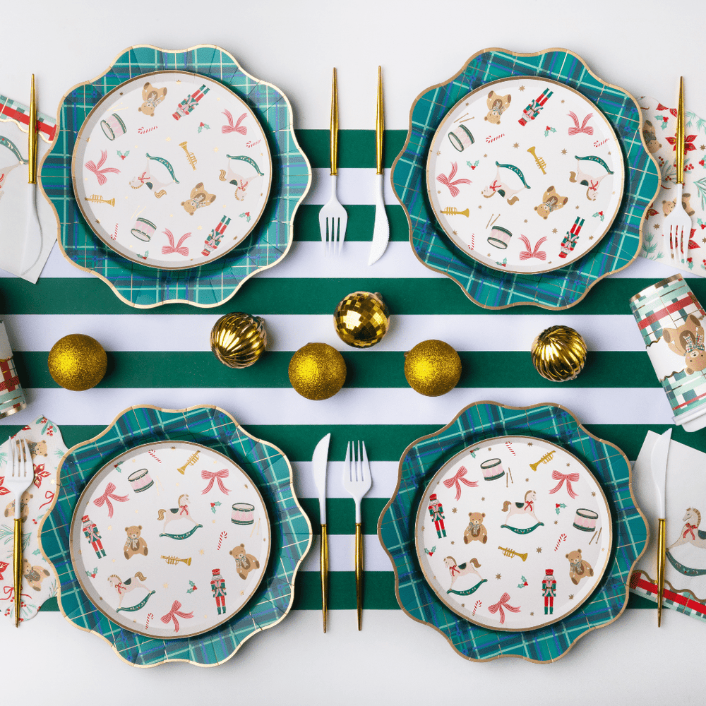 Four holiday-themed dinnerware plates arranged in a flat lay on a white and green striped table runner, with gold flatware, gold ornaments, and patterned napkins around them.