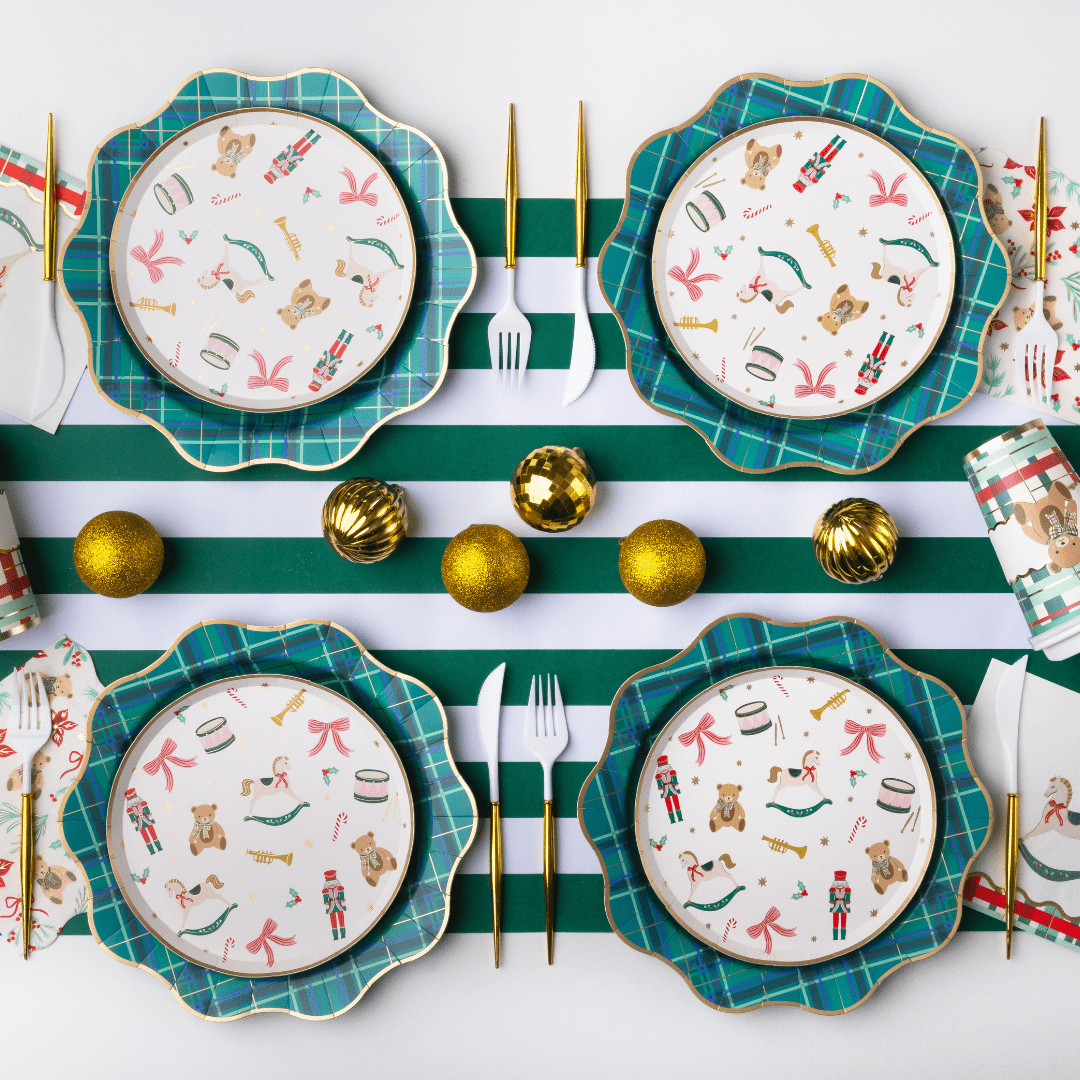 Four holiday-themed dinnerware plates arranged in a flat lay on a white and green striped table runner, with gold flatware, gold ornaments, and patterned napkins around them.