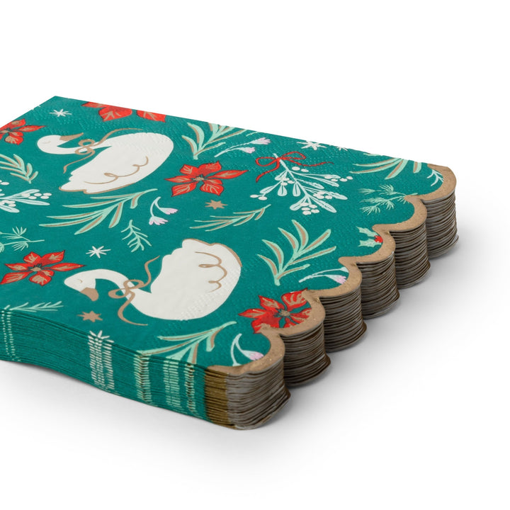 side angle of a paper napkin with a scalloped gold edge, featuring a festive holiday pattern of white swans, red poinsettias, mistletoe, and gold stars on a teal background, isolated on a white studio background.