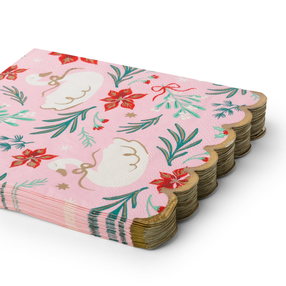 A stack of pink paper napkins with scalloped edges, featuring a festive holiday pattern of white swans, red poinsettias, green mistletoe, and gold stars, displayed on a clean white background.