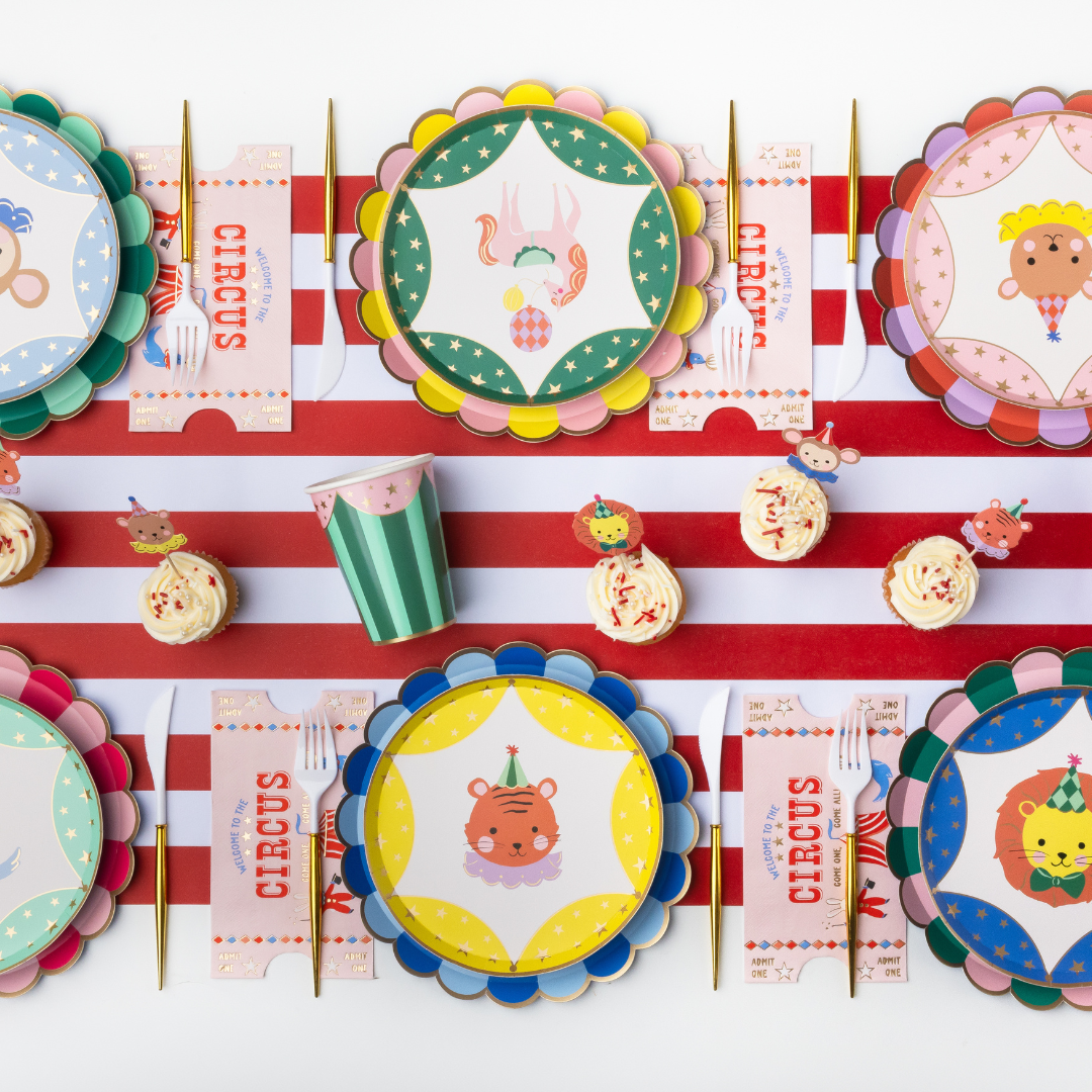 A set of circus-themed party tableware arranged in a flat lay on a red and white striped background. The set includes scalloped paper plates, matching napkins, gold-colored forks and knives, a striped paper cup, and small cupcakes with decorative toppers.