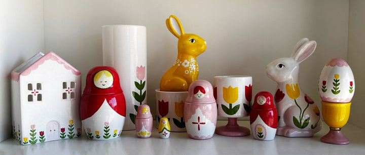 Red and yellow tulip cup on a shelf with a ceramic house, nesting dolls, and golden and white bunny figures