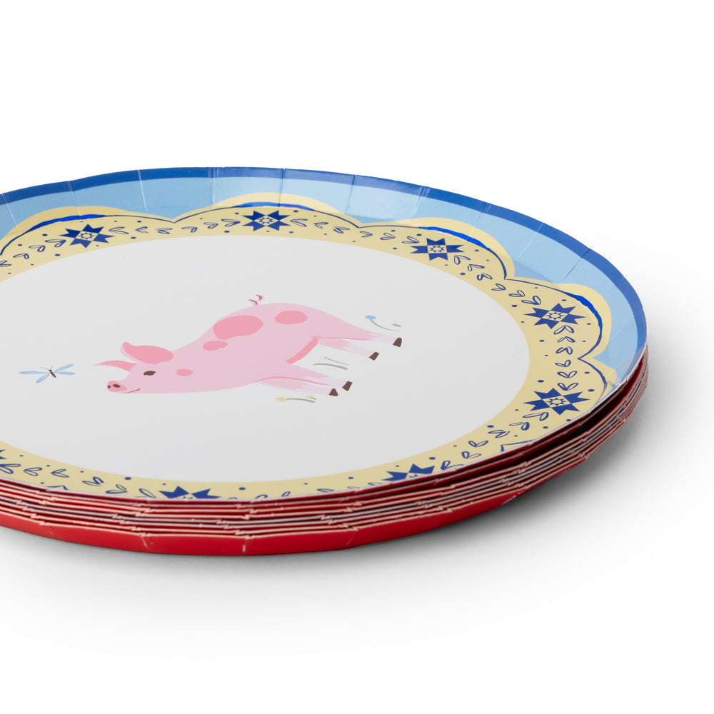 A stack of round paper plates featuring a whimsical pink pig illustration in the center, surrounded by a scalloped cream border with blue floral motifs and a blue rim, with red rims visible on the stacked plates.