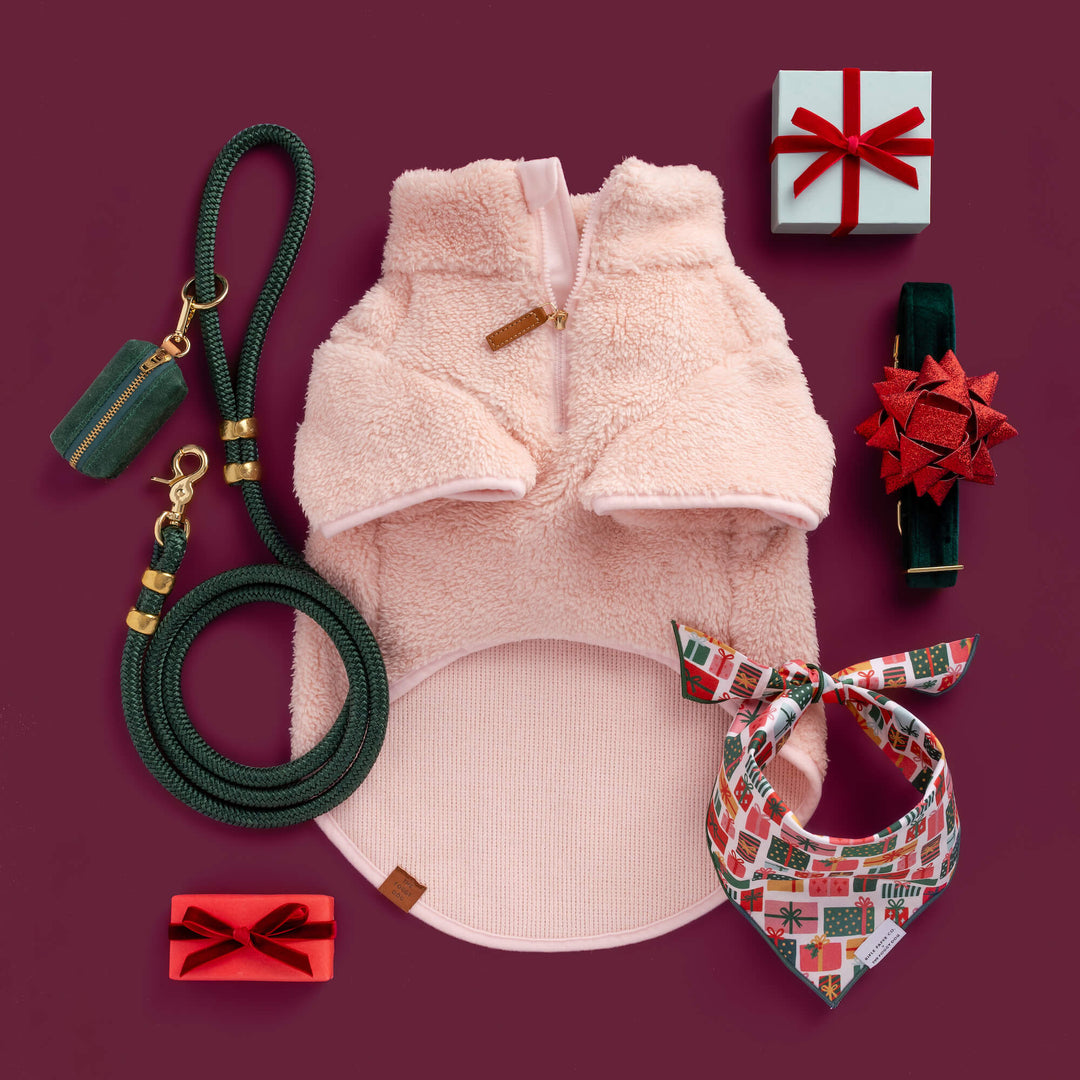 Holiday dog gear on burgundy: pink fleece, green leash and bag holder, gift-print bandana, and red-bow green collar.
