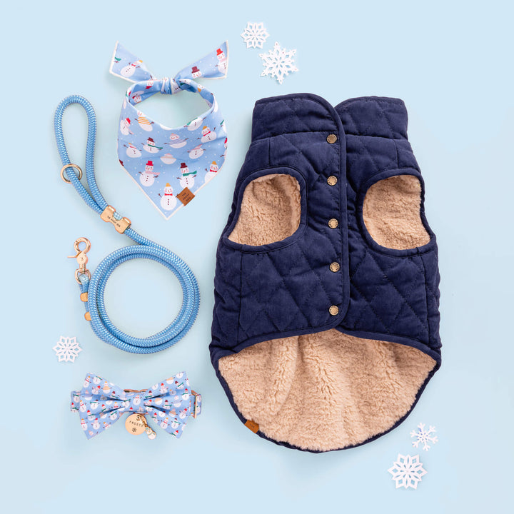 Winter flat lay on light blue with a navy quilted dog vest, blue rope leash, and snowman print bandana
