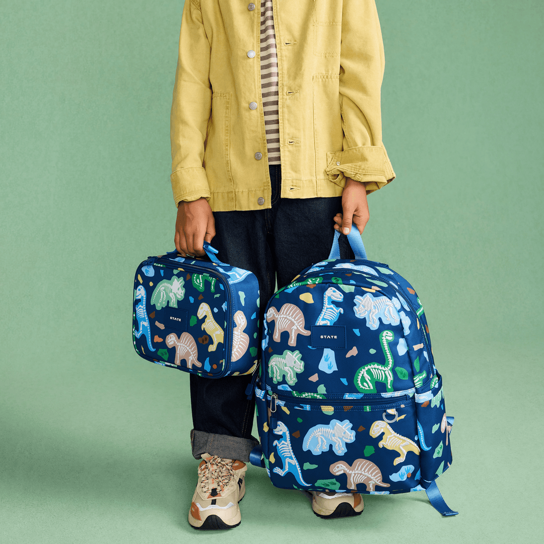 Child holding the Dino Fossils lunch box beside the matching Dino Fossils Kane Backpack