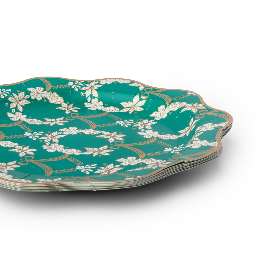 Side angle of scalloped paper plate featuring a teal green background with a repeating white floral wreath pattern and a metallic gold border.