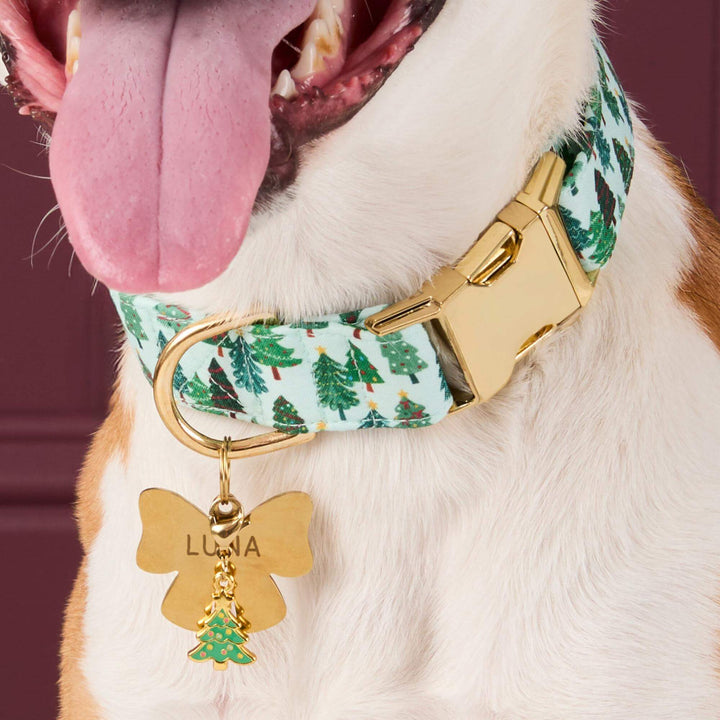 Christmas tree collar charm beneath a gold bow-shaped name tag on a dog's pale green tree-print collar.