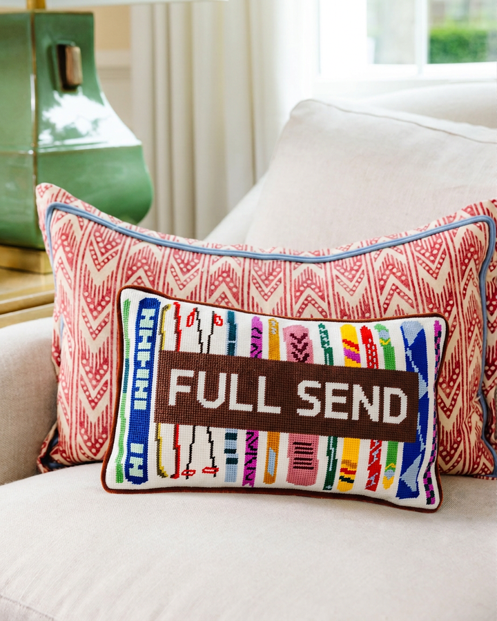 Full Send ski needlepoint pillow styled on a cream sofa with a red patterned pillow for a ski lodge living room