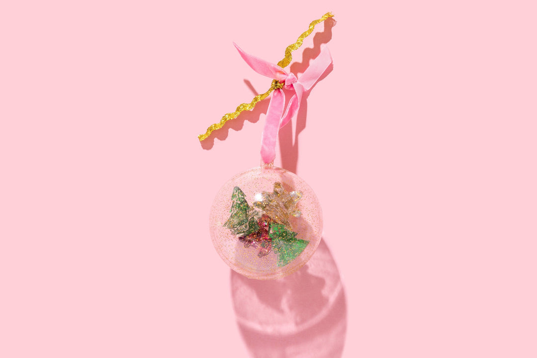 Clear glitter ball ornament packaging with tree clips inside, tied with a pink ribbon on a pink backdrop