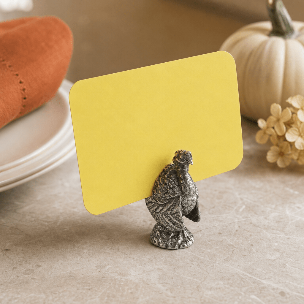 Pewter turkey place card holder with a yellow card beside white plates and a white pumpkin