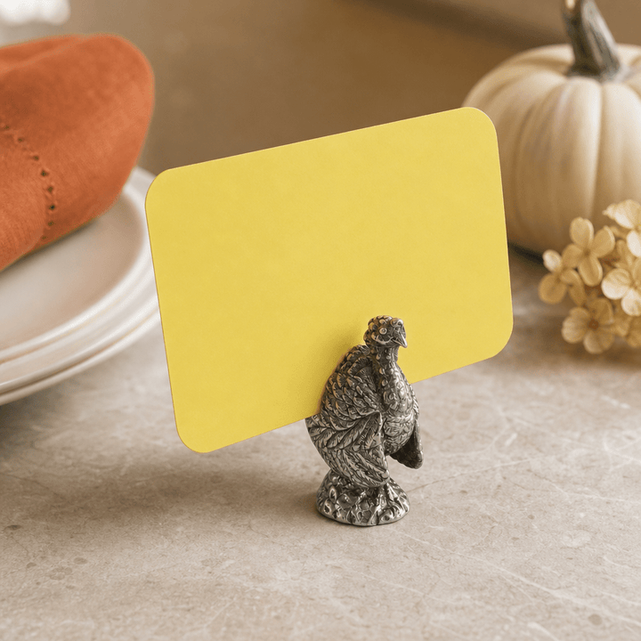Pewter turkey place card holder with a yellow card beside white plates and a white pumpkin
