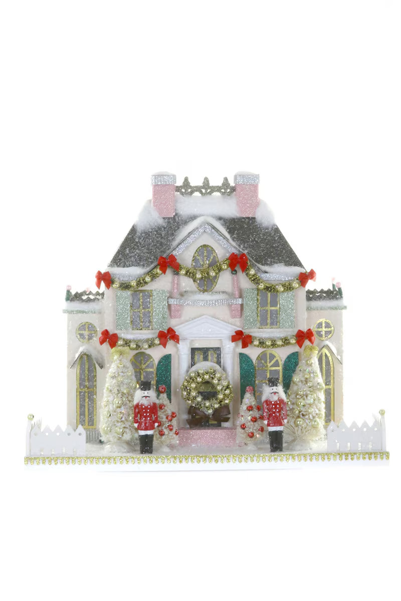 White glitter paper Christmas mansion with nutcracker guards, a wreath, and bottle brush trees for a holiday village