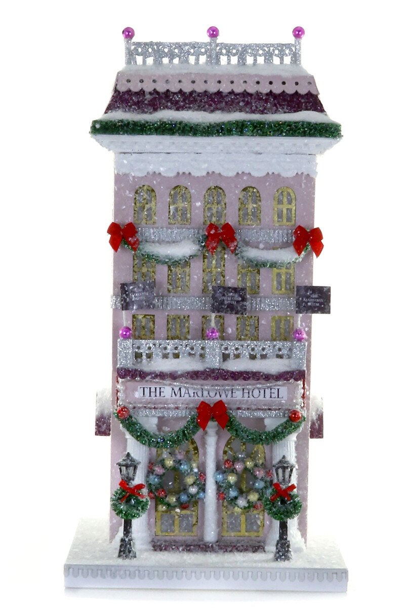 Pink glitter paper Marlowe Hotel Christmas village building with wreaths, garlands, red bows, and bottle brush trees for holiday mantel decor