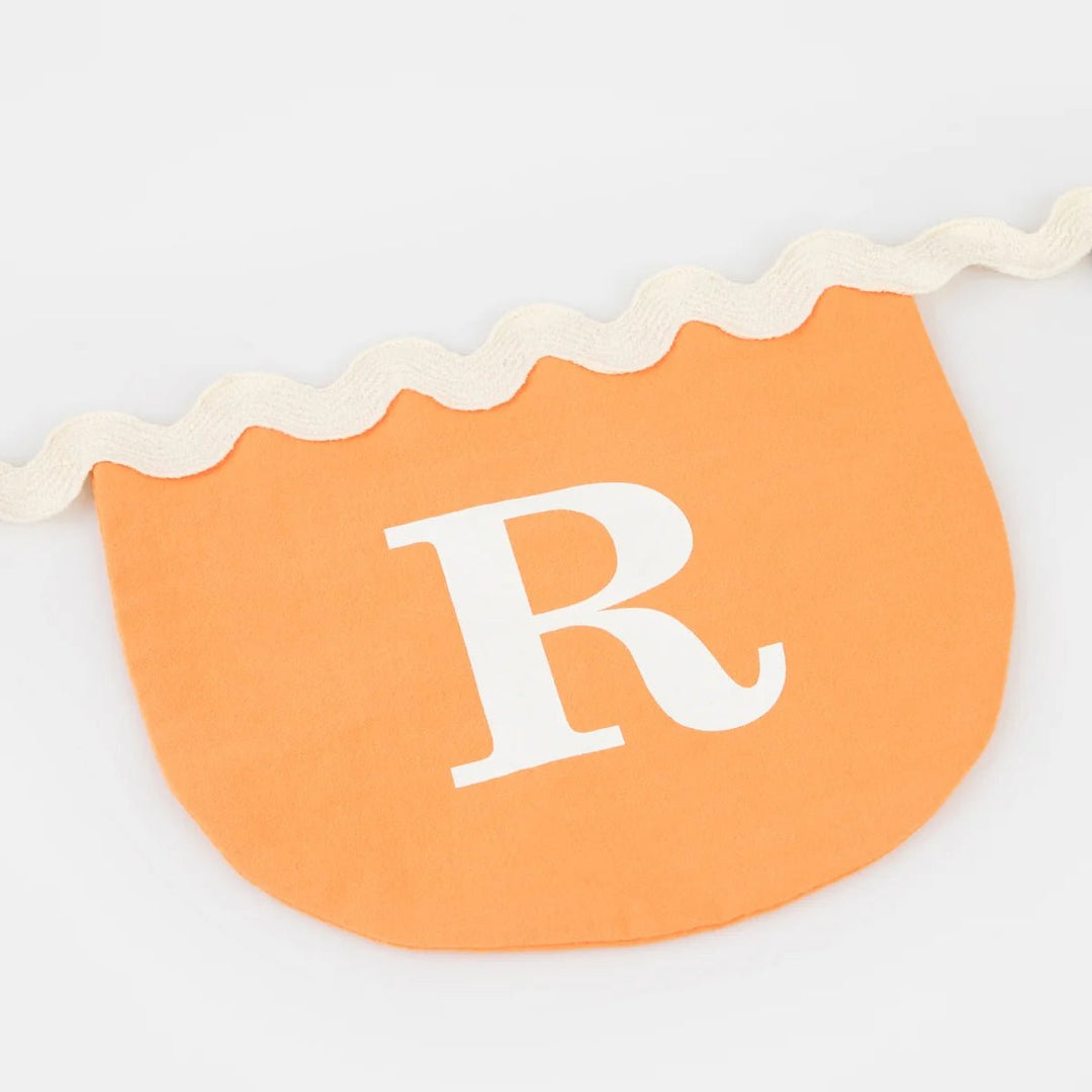 Close view of a scalloped orange fabric pennant with a white letter R and cream ric rac trim on the garland cord