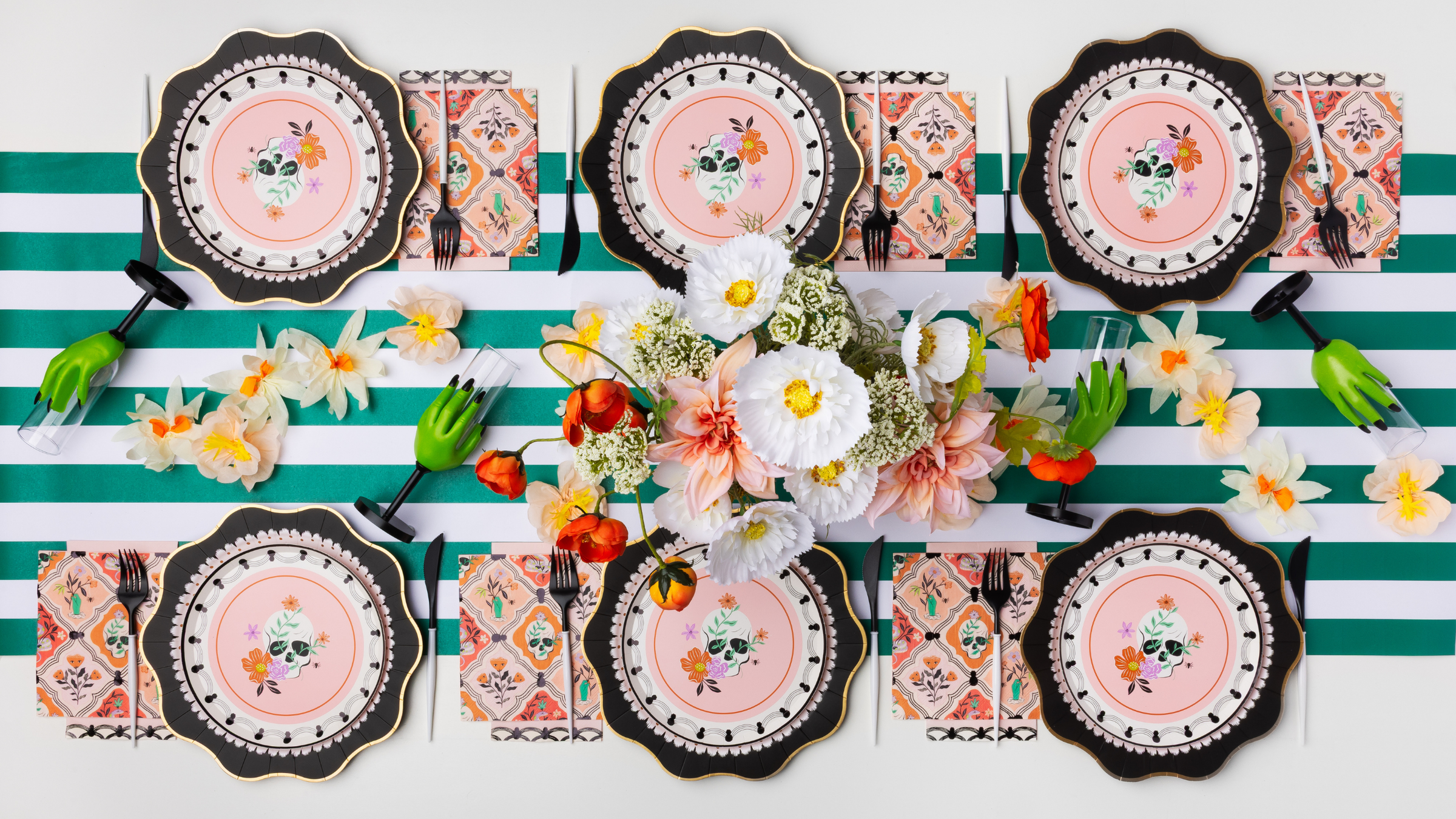 A styled table setting featuring Day of the Dead themed place settings with pink sugar skull plates, patterned napkins, and a central floral centerpiece on a green and white striped background.