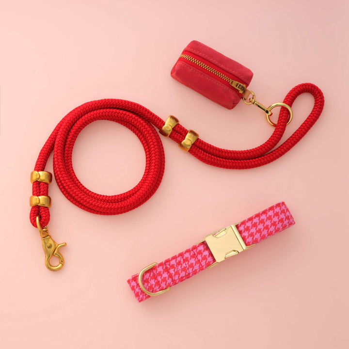 Houndstooth collar, red rope leash, and red bag holder arranged on a blush pink backdrop