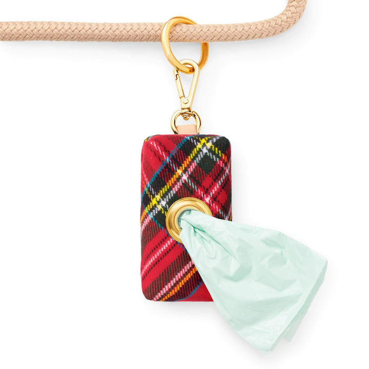 Red tartan waste bag holder clipped to a cream leash, dispensing a pale green bag through its brass eyelet.