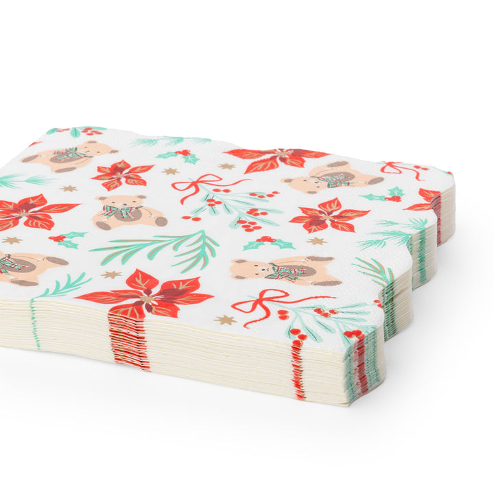 A stack of white paper napkins featuring a festive Christmas pattern with teddy bears, red poinsettias, green pine branches, holly leaves, and red bows, shown on a clean white background.