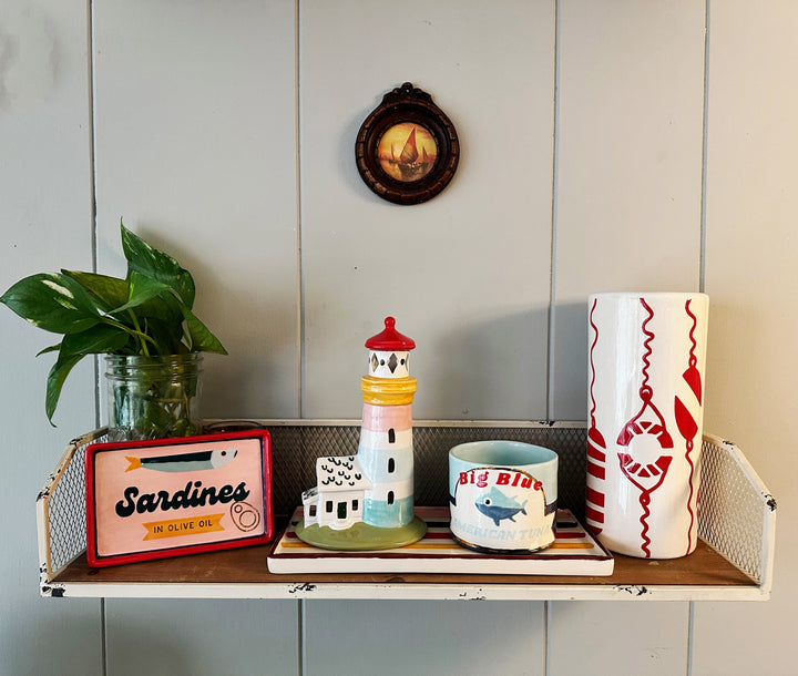 Decorative shelf with various items including a lighthouse, 'Sardines' sign, and ceramic mug.
